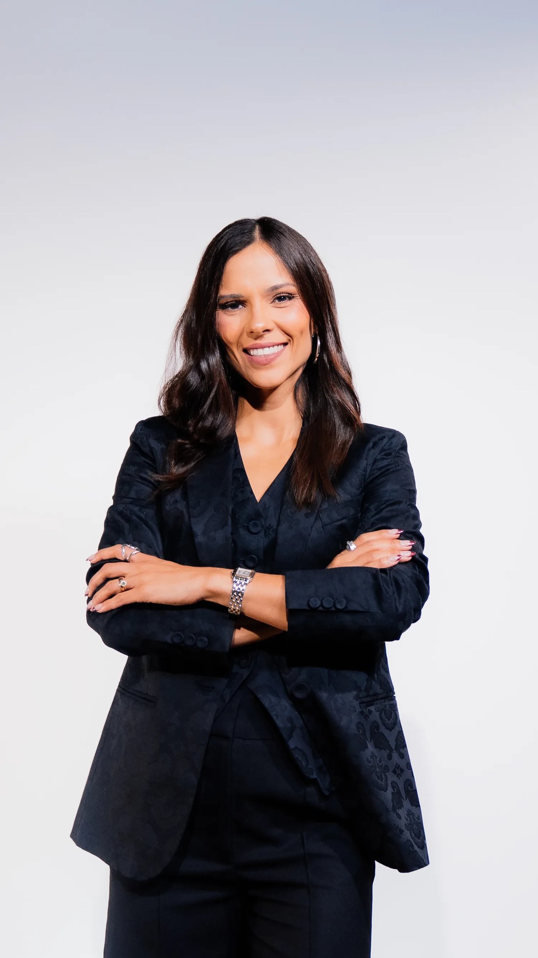 Ana Gabriela Fidalgo, de traje negro y brazos cruzados, sonriendo a cámara sobre fondo claro de estudio.