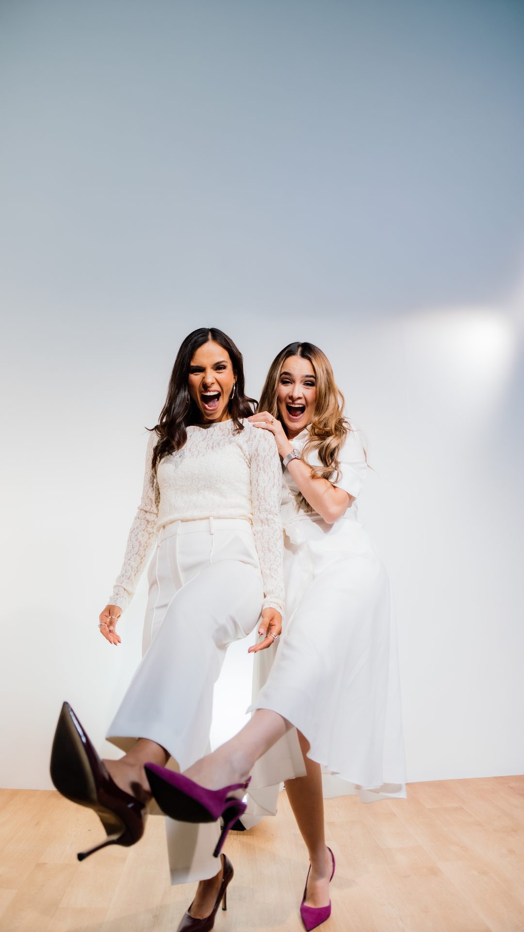 Michelle Ruiz y Ana Gabriela Fidalgo, vestidas de blanco, de cuerpo entero y riendo en un estudio de fondo claro.